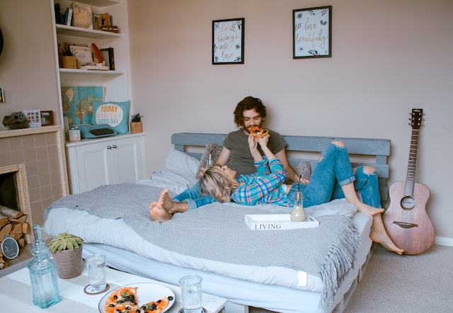 una pareja acostada en una cama mientras come pizza y una guitarra a su lado